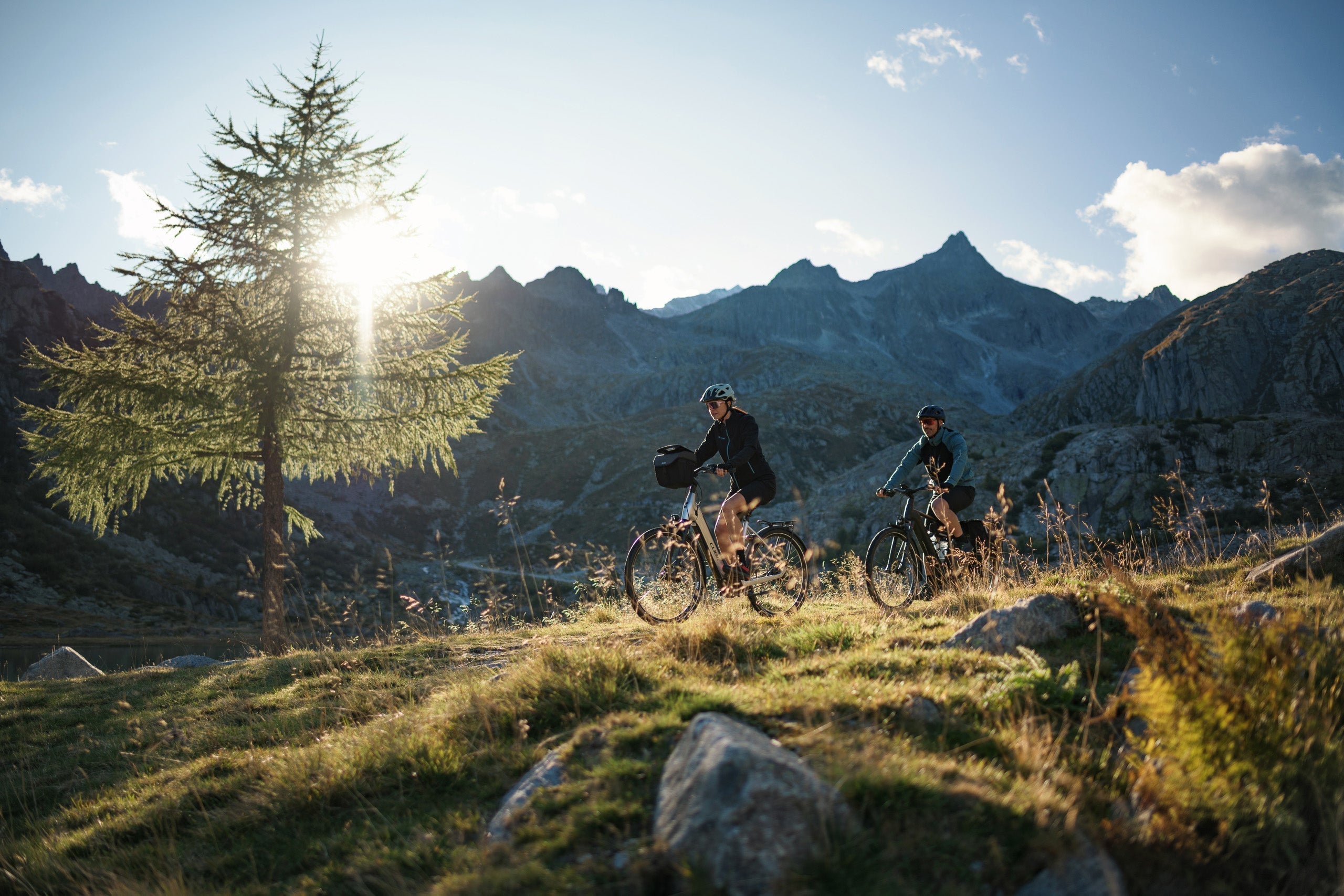 Zwei Personen fahren bei Sonnenuntergang auf einem grasbewachsenen Hügel Fahrrad, mit einem Baum in der Nähe und schroffen Bergen im Hintergrund. Die Sonne scheint durch den Baum und wirft ein warmes Licht auf die malerische Landschaft.