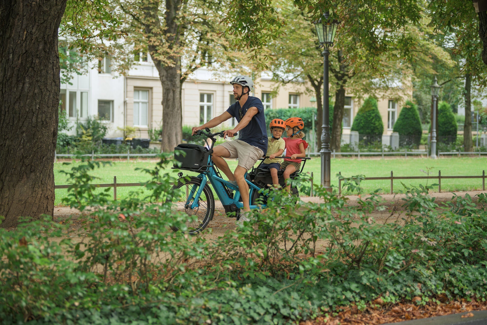Ein Mann fährt auf einem Fahrrad, auf dem zwei Kinder mit Helmen sitzen, durch einen Park mit Bäumen und Grünanlagen.