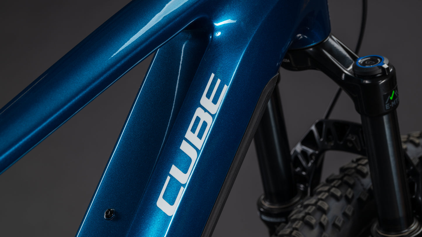 Nahaufnahme eines CUBE Stereo Hybrid ONE22 Race 800 electricblue'n'white 2026 Mountainbike-Rahmens, mit dem weißen CUBE-Logo und einem Teil der vorderen Federgabel mit einem Noppenreifen im Hintergrund.