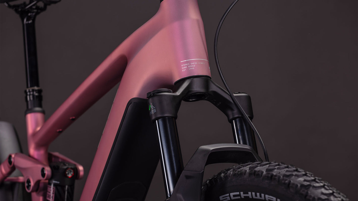 Nahaufnahme des CUBE Stereo Hybrid ONE22 Race 800 shiftblush'n'black 2026 E-Mountainbikes. Hervorzuheben sind die mattrosa Lackierung, die Federgabel und die Schwalbe-Reifen. Der Shimano XT-Antrieb sorgt für reibungslose Schaltvorgänge vor einem dunklen Hintergrund.