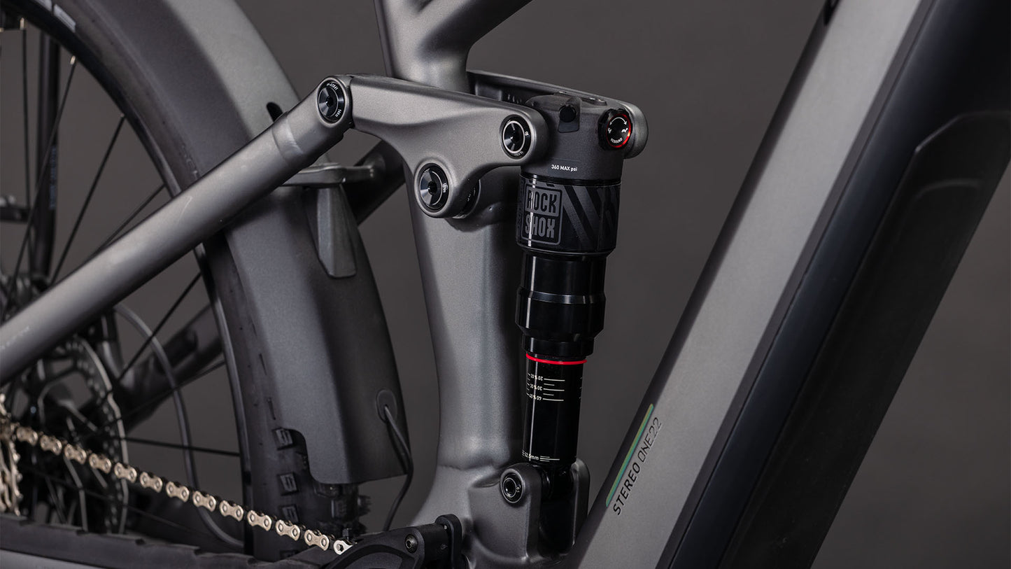 Nahaufnahme des CUBE Stereo Hybrid ONE22 Race 800 FE slabgrey'n'chrome 2026 Rahmens, die das mattgraue Finish, die Hinterradfederung mit RockShox Dämpfer und die sichtbare Kette in der Nähe des Hinterrads zeigt.