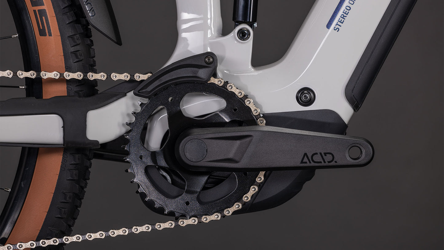 Nahaufnahme des CUBE Stereo Hybrid ONE22 SLX 800 greige'n'switch 2026 mit ACID-Kurbelarm, Kettenblatt, Fahrradkette, Shimano XT-Schaltung und Hinterradreifen. Der weiß-schwarze Rahmen hebt sich von einem dunklen Hintergrund mit scharfen Details ab.