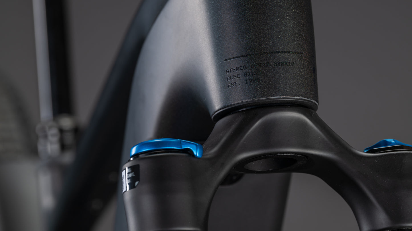 Nahaufnahme des CUBE Stereo Hybrid ONE22 SLT 800 prizmblack'n'black 2026 Rahmen und Gabel in mattem Schwarz, mit blauen Einstellknöpfen und einem dezent eingravierten Etikett, vor einem verschwommenen Hintergrund.