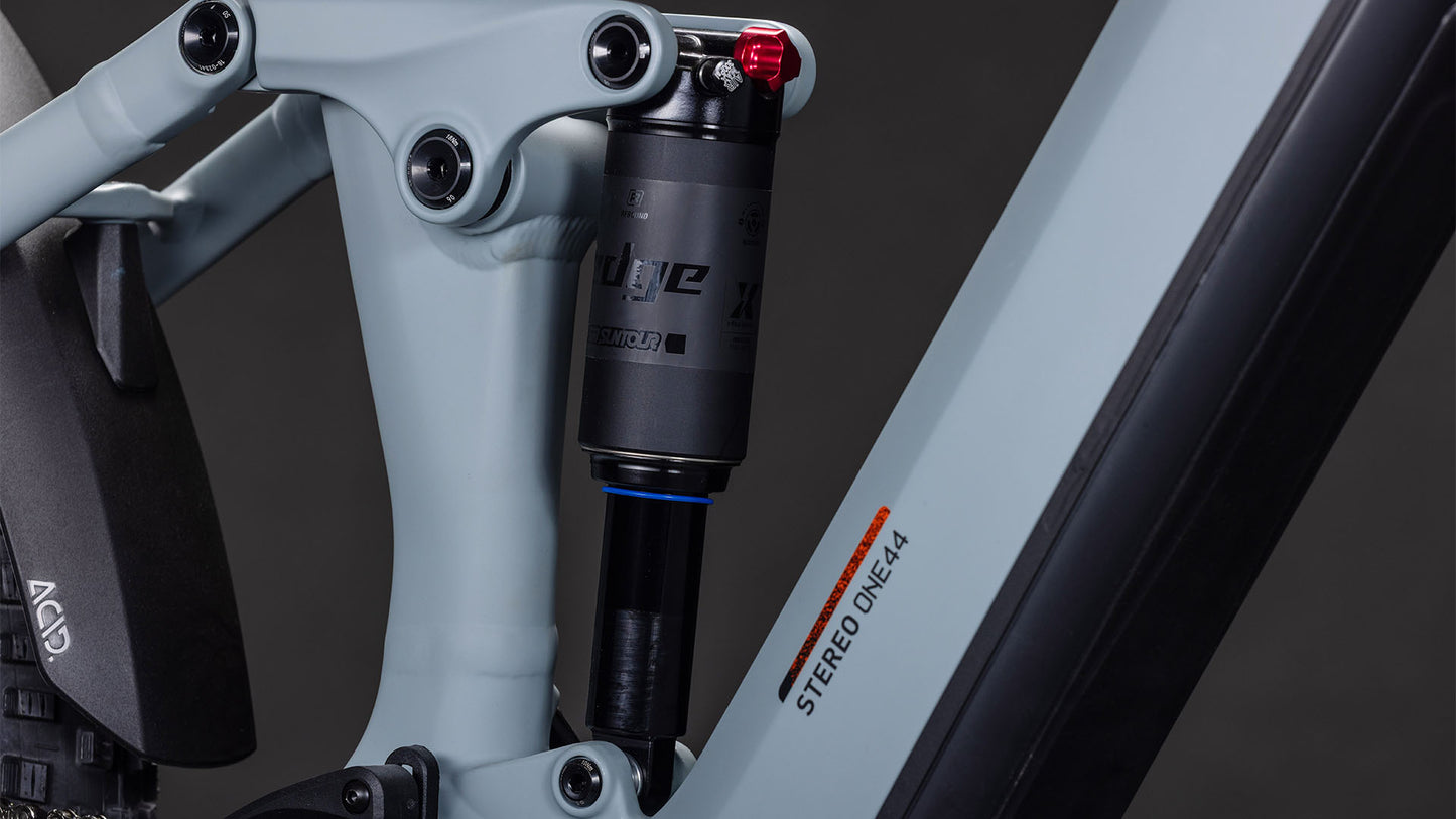 Nahaufnahme der Hinterradfederung des CUBE Stereo Hybrid ONE44 Pro 800. Zu sehen sind der Stoßdämpfer, der Rahmen mit der Aufschrift "STEREO ONE44" und die Shimano XT-Schaltung in Hellgrau und Orange vor dunklem Hintergrund.