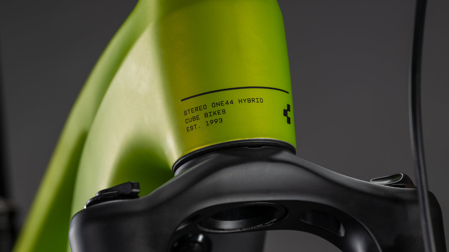 Nahaufnahme eines Cube Stereo Hybrid ONE44 Pro 800 lizard'n'black Rahmens über einer schwarzen Vorderrad-Federgabel mit der Aufschrift "STEREO ONE44 HYBRID CUBE BIKES EST. 1993" auf dem Rahmen vor einem dunklen, unscharfen Hintergrund.