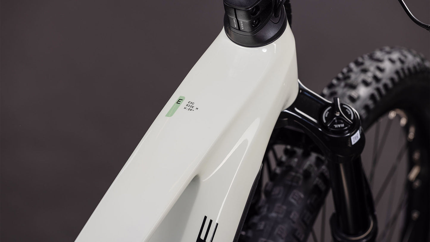 Nahaufnahme des Vorderrahmens eines CUBE Stereo Hybrid ONE44 EXC 800 desertstone'n'driedherbs 2026 E-Bikes mit Gabel, Laufrad mit dickem Profil und einem Teil des Lenkers vor einem dunklen Hintergrund, mit dezentem Branding und einem kleinen Display am Rahmen.