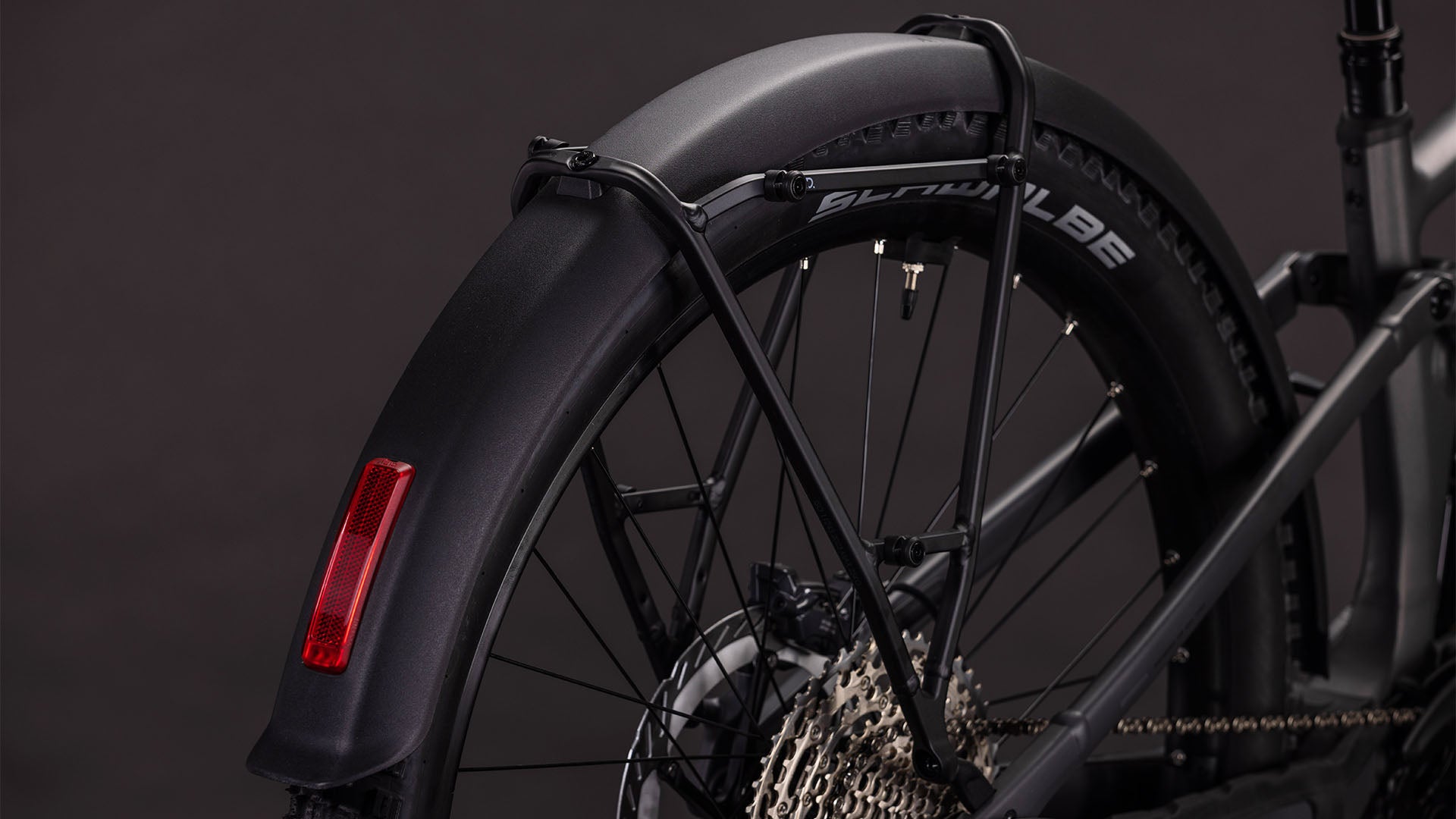 Nahaufnahme des Hinterrads des CUBE Stereo Hybrid ONE44 EXC 800 FE irongrey'n'chrome 2026, wobei ein schwarzes Schutzblech mit rotem Reflektor, eine Shimano XT-Kassette und ein Schwalbe-Reifen vor einem dunklen Hintergrund hervorgehoben werden.