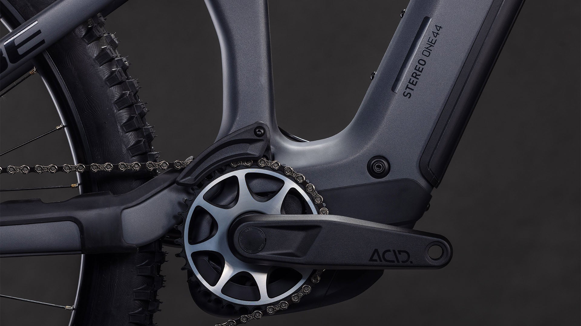 Nahaufnahme der CUBE Stereo Hybrid ONE44 HPC SLX 800 slabgrey'n'orange 2026 Kurbelgarnitur und Kette, mit Shimano XT Schaltung und Bosch CX Motor auf dem schlanken Rahmen mit STEREO ONE44 und ACID Aufschriften vor einem dunklen Hintergrund.