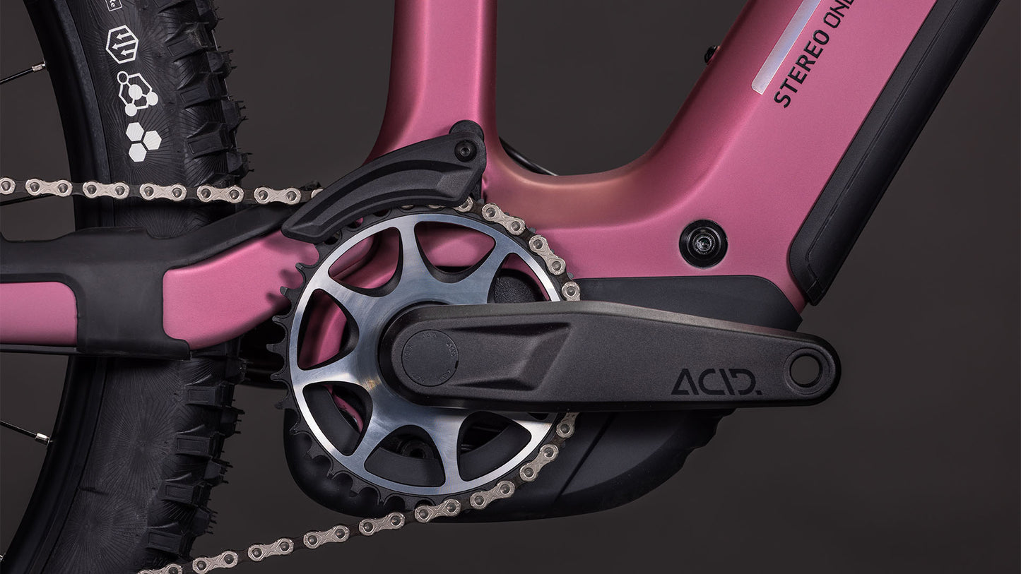 Nahaufnahme der CUBE Stereo Hybrid ONE44 HPC SLX 800 shiftblush'n'art 2026 Kurbelgarnitur, des ACID-gekennzeichneten Pedalarms und des Kettenblatts, wobei ein Teil des Hinterradreifens und des Rahmenlogos zu sehen ist, was die fortschrittliche Integration des Bosch CX-Motors zeigt.