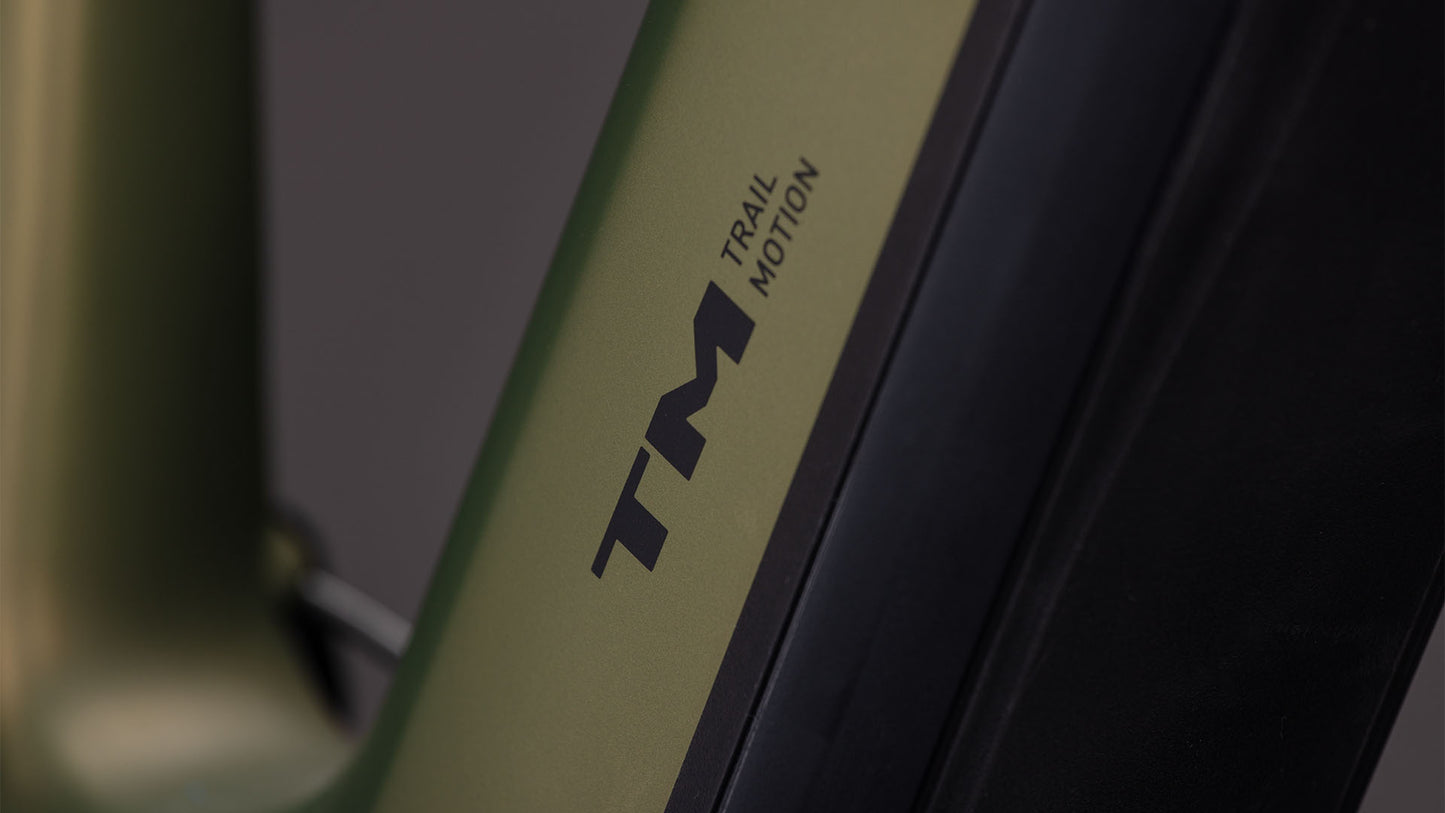 Nahaufnahme eines CUBE Stereo Hybrid ONE44 HPC TM 800 reedgreen'n'matrix 2026 E-Mountainbike mit mattgrünem und schwarzem Rahmen, auf dem der Text TM TRAIL MOTION in schwarzer Schrift zu lesen ist. Der Hintergrund ist unscharf und grau.