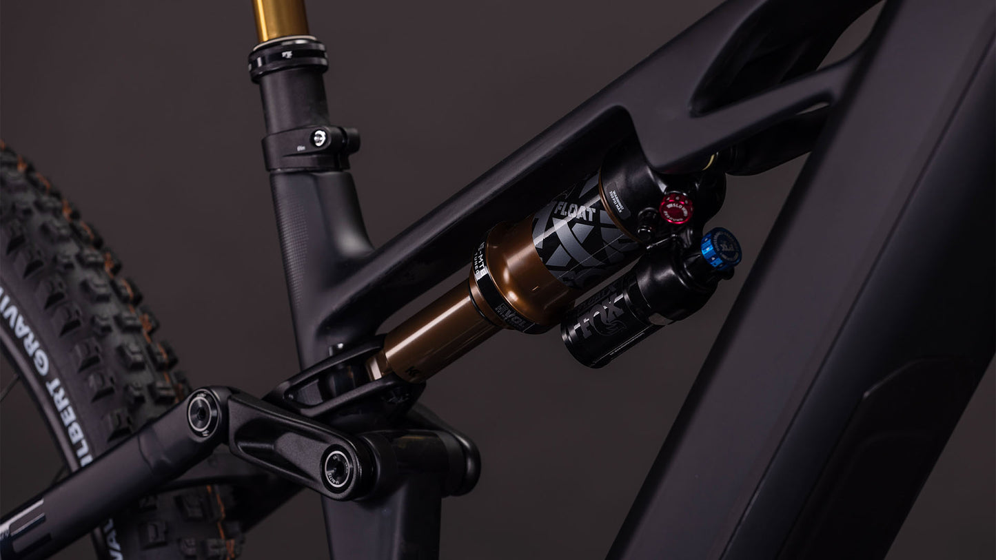 Nahaufnahme der Hinterradfederung des CUBE Stereo Hybrid ONE44 HPC AT 800 carbon'n'gold 2026 mit einem FOX FLOAT X-Dämpfer auf einem mattschwarzen Rahmen. Shimano XT-Komponenten steigern die Leistung, ein Teil des Hinterreifens und des Sitzrohrs sind sichtbar.
