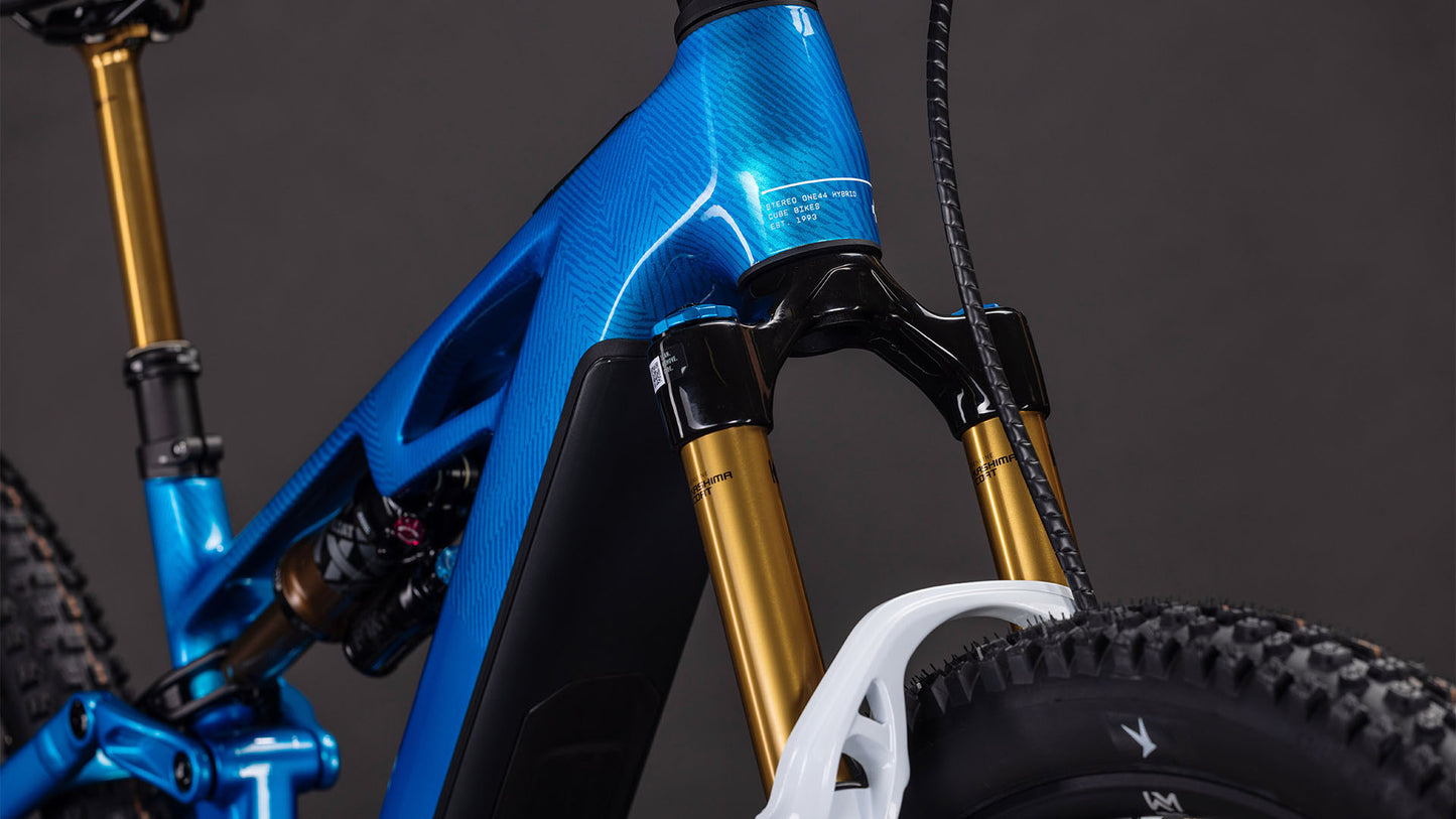 Nahaufnahme des CUBE Stereo Hybrid ONE44 HPC AT 800 actionteam 2026 E-Bike Rahmens in Blau, Federgabel mit goldenen Standrohren, Shimano XT Komponenten und Reifenprofil vor dunklem Hintergrund.