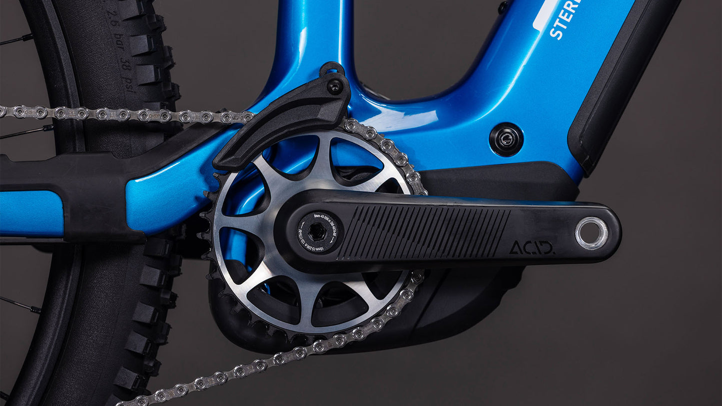 Nahaufnahme der CUBE Stereo Hybrid ONE44 HPC AT 800 actionteam 2026 Kurbelgarnitur, des Kettenblatts und der Kette mit Pedal und Hinterradreifen vor einem dunklen Hintergrund, der die detaillierten Schaltkomponenten zeigt.