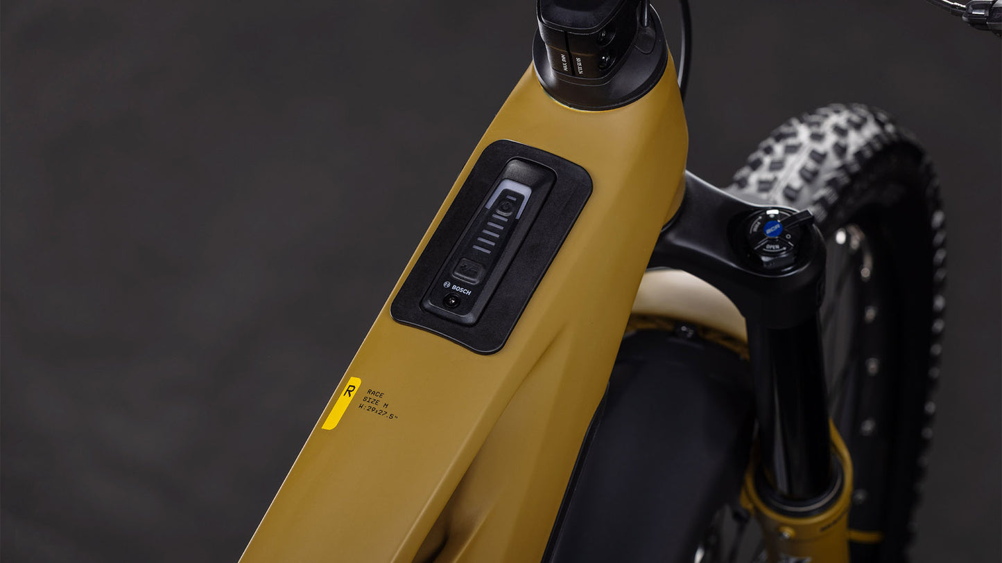 Nahaufnahme des Bedienfelds am Oberrohr des CUBE Stereo Hybrid ONE77 HPC Race 800 willowgreen'n'black 2026 E-Mountainbikes. Zu sehen sind Tasten, eine LED-Anzeige, Teile des Lenkers und der Hinterreifen.