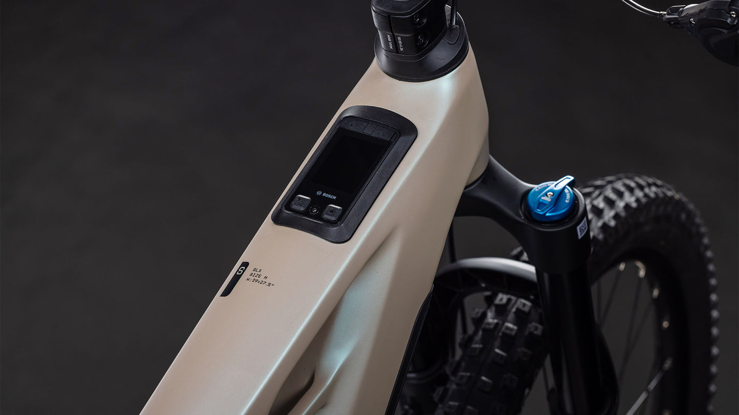 Nahaufnahme des CUBE Stereo Hybrid ONE77 HPC SLX 800 in glintsand'n'black: beiger E-Mountainbike-Rahmen mit integriertem Display, Bedientasten am Oberrohr, Bosch CX-Motor, Noppen-Vorderreifen und Fox Float 38 Rhythm-Federgabel.