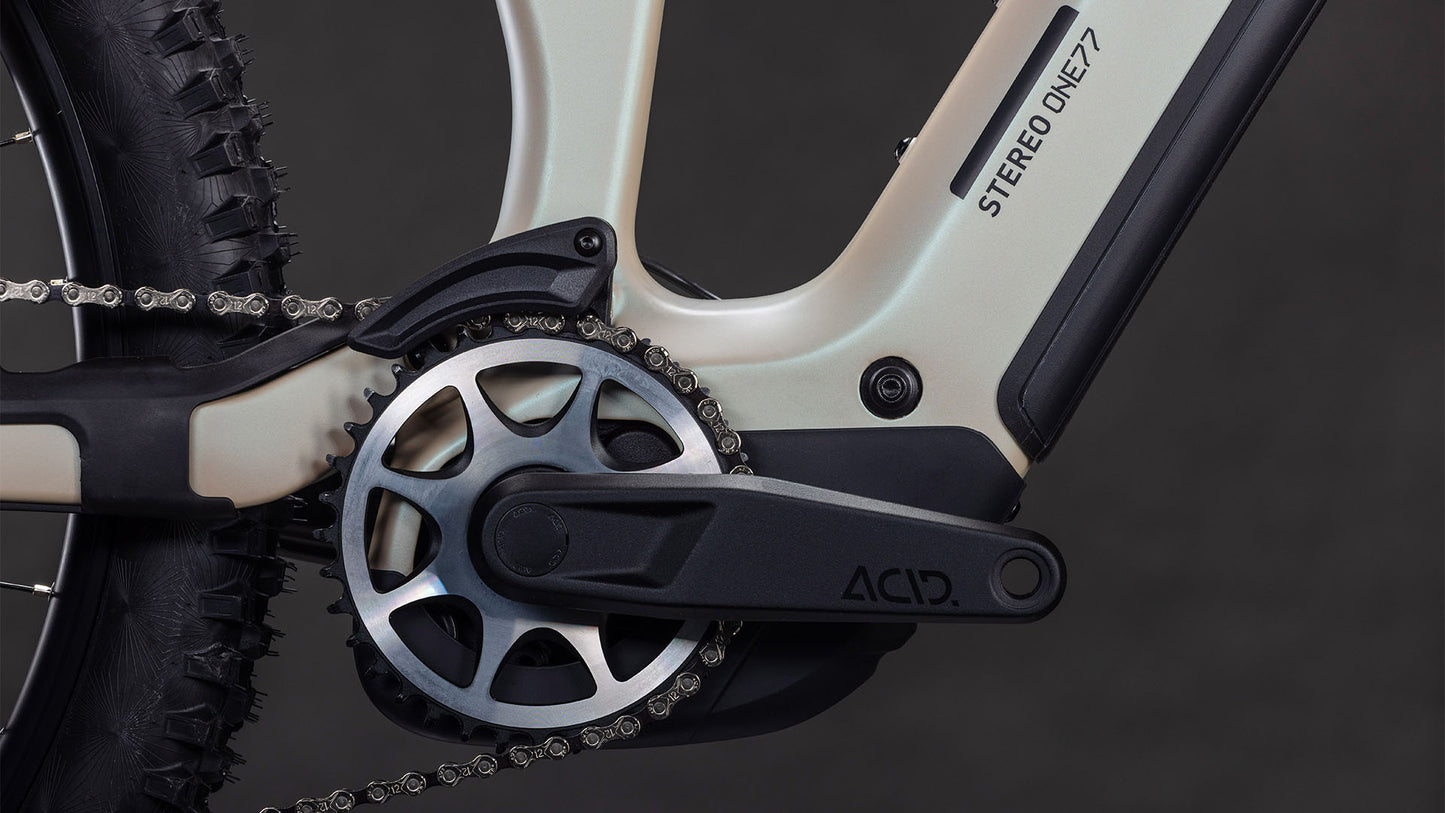 Nahaufnahme einer CUBE Stereo Hybrid ONE77 HPC SLX 800 Kurbelgarnitur, Kettenblatt und ACID Pedalarm am glintsand'n'black Rahmen. Das Bild zeigt auch den knorrigen Hinterreifen, das Kettensegment und die Shimano XT Schaltung.