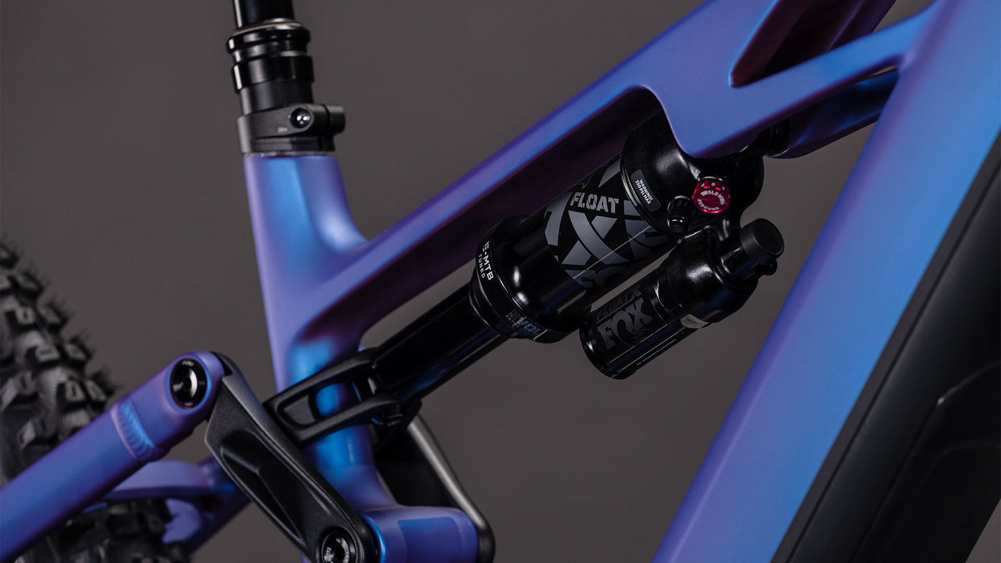Nahaufnahme der Hinterradfederung des CUBE Stereo Hybrid ONE77 HPC SLX 800 Mountainbikes mit FOX FLOAT Stoßdämpfer und markantem blau-iris'n'black Rahmen vor dunklem Hintergrund.