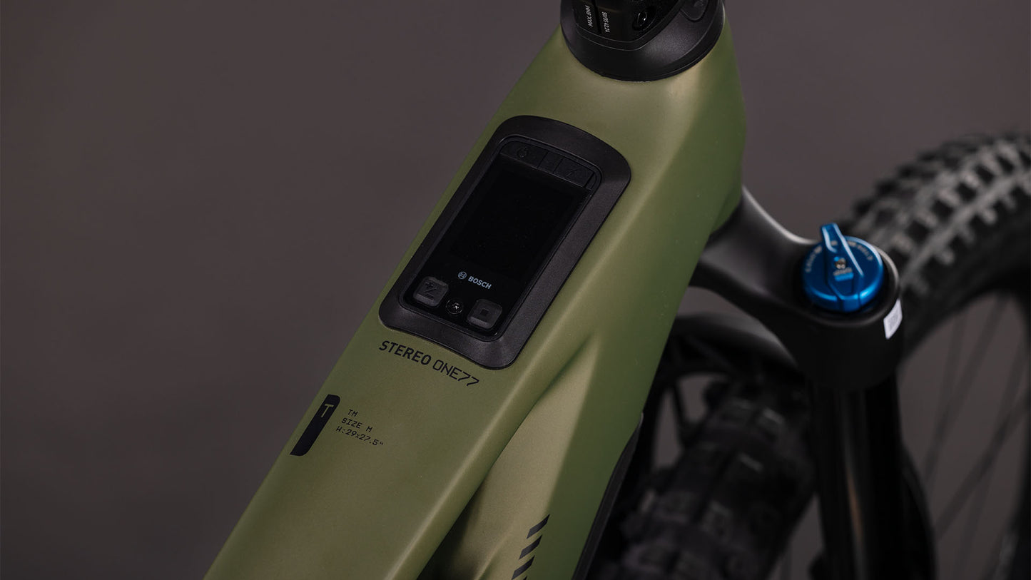 Nahaufnahme des Oberrohrs des CUBE Stereo Hybrid ONE77 HPC TM 800 (reedgreen'n'matrix, 2026) mit eingebauter Bosch CX-Motoranzeige und Bedienfeld. Der mattgrüne Rahmen mit der Aufschrift "STEREO ONE77", mit Hinterradreifen, blauer Gabel und 170 mm Federweg ist teilweise sichtbar.
