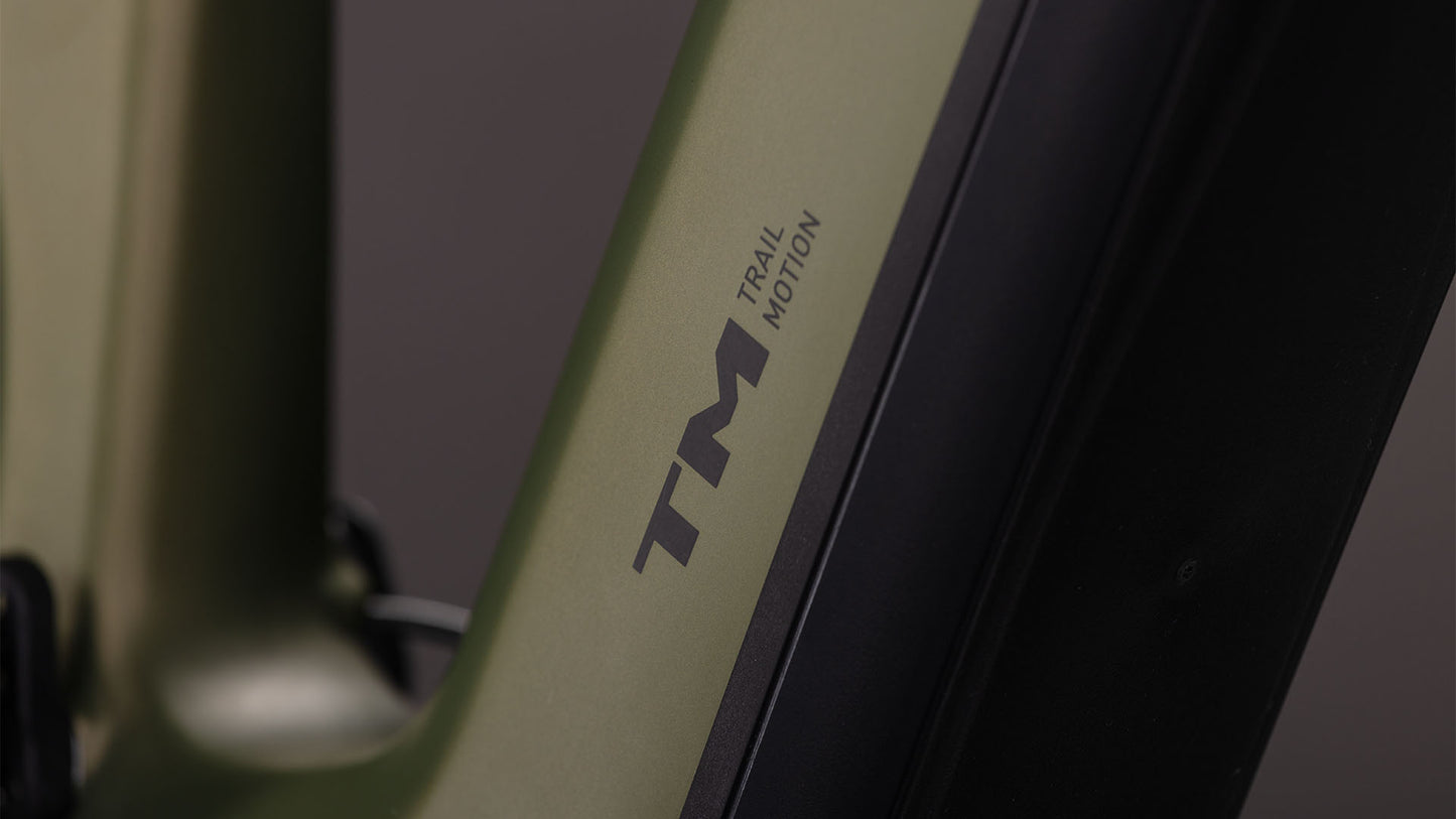 Nahaufnahme des CUBE Stereo Hybrid ONE77 HPC TM 800 reedgreen'n'matrix 2026 Rahmens mit "TM" und "Trail Motion" in schwarz, neben einer schwarzen Sram GX Eagle Getriebekomponente. Der Hintergrund ist unscharf.
