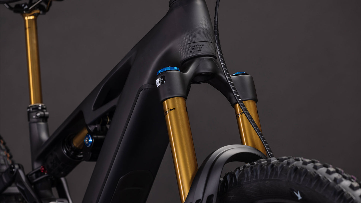 Nahaufnahme des CUBE Stereo Hybrid ONE77 HPC AT 800 carbon'n'gold 2026, die den mattschwarzen Carbonrahmen, die goldene Federgabel mit blauen Einstellknöpfen und den Vorderreifen vor dunklem Hintergrund zeigt - gebaut für Big-Mountain-Riding.