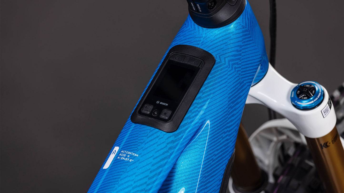 Die Nahaufnahme des Oberrohrs des CUBE Stereo Hybrid ONE77 HPC AT 800 actionteam 2026 Elektrofahrrads zeigt den blau gemusterten Rahmen, das Display des Bosch CX-Motors und die Bedienknöpfe in der Nähe des Lenkervorbaus.