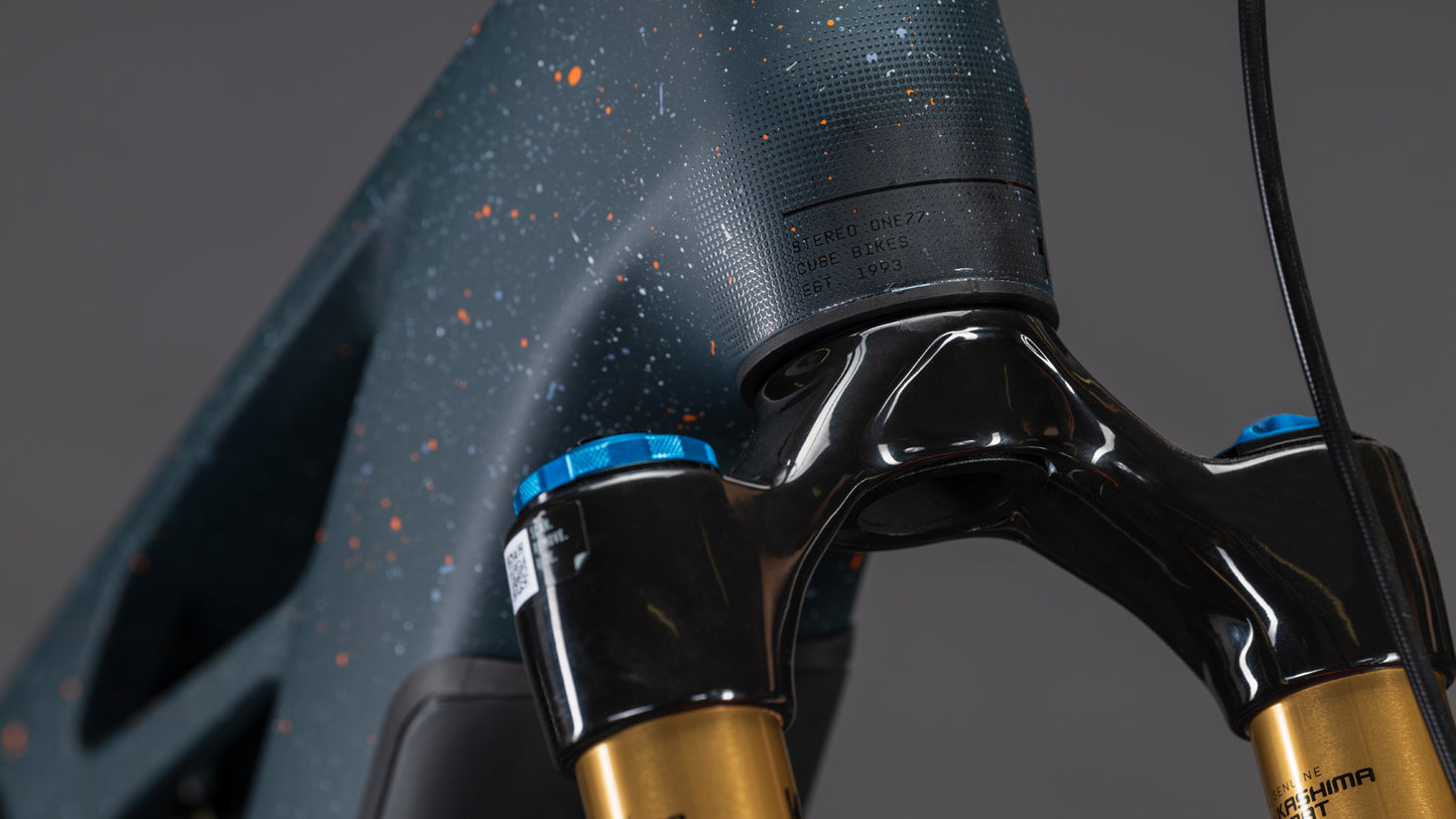 Nahaufnahme des CUBE Stereo Hybrid ONE77 HPC SLT 800 E-Bike in galaxy'n'orange (2026), mit goldener und blauer Federgabel und leistungsstarkem Bosch CX-R Motor, vor einem schlichten grauen Hintergrund.