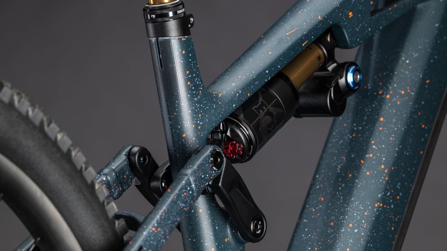 Nahaufnahme der Hinterradaufhängung und des Rahmens des CUBE Stereo Hybrid ONE77 HPC SLT 800 galaxy'n'orange 2026 in mattem Blau mit orangefarbenen Sprenkeln, die den Stoßdämpfer, die Drehpunkte und die Big-Mountain-Performance in der Nähe des Hinterrads hervorheben.