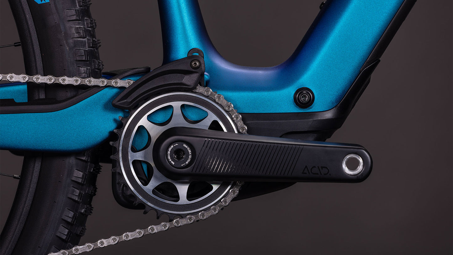 Nahaufnahme der Kurbelgarnitur und des Kettenblatts des CUBE AMS Hybrid ONE44 C:68X SLX 400X nebula'n'black 2026, mit der Kette, dem mit ACID beschrifteten Pedalarm und einem Teil des Hinterradreifens vor einem dunklen Hintergrund.