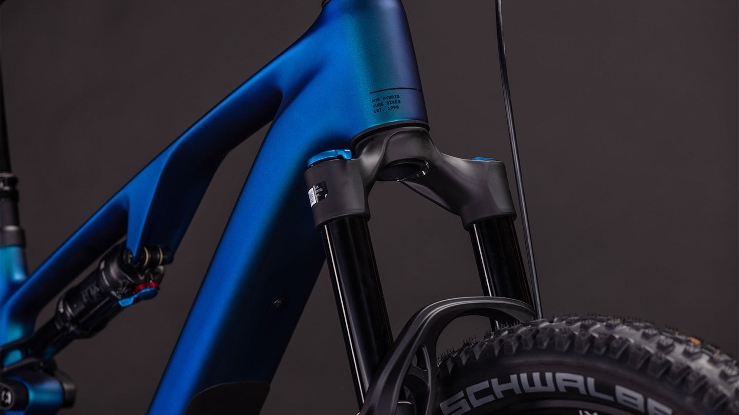 Nahaufnahme des CUBE AMS Hybrid ONE44 C:68X SLX 400X nebula'n'black 2026, die den blauen C:68X-Carbonrahmen, die Federgabel, den Stoßdämpfer und die Schwalbe-Reifen vor einem dunklen Hintergrund hervorhebt.