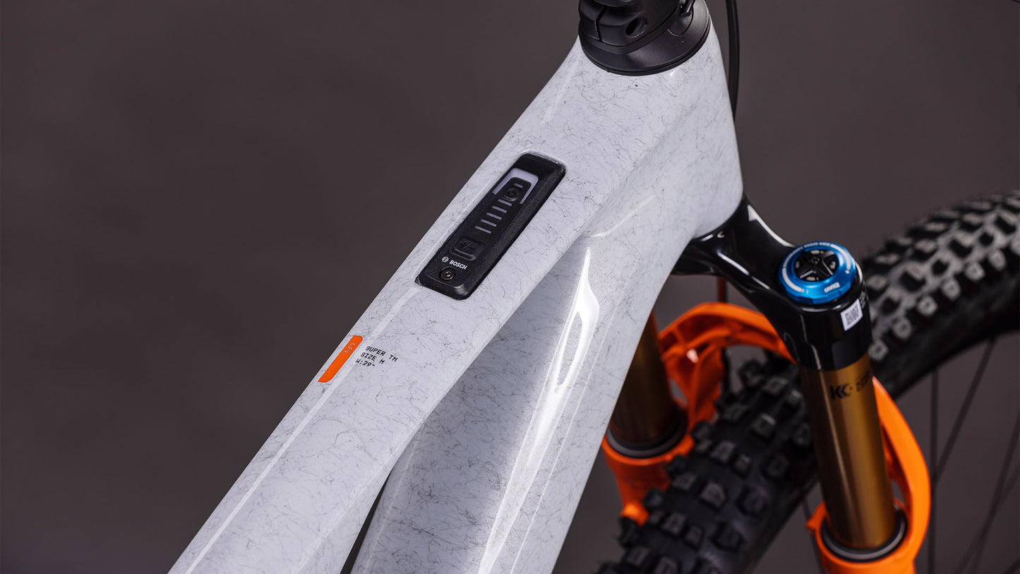 Nahaufnahme eines Cube AMS Hybrid ONE44 C:68X SUPER TM 400X von CUBE in foggrey'n'fibre, die den Carbonrahmen mit elektronischem Bedienfeld, die orangefarbene Federgabel, das matte, strukturierte Finish und die Details des Vorderreifens zeigt.