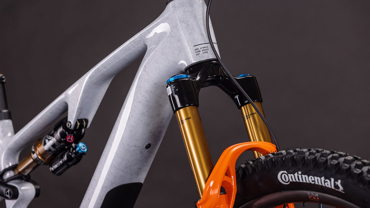 Nahaufnahme des CUBE AMS Hybrid ONE44 C:68X SUPER TM 400X foggrey'n'fibre 2026 Rahmens mit goldener Federgabel, orangefarbenem Gabelbogen, hinterem Stoßdämpfer und Continental Reifen vor dunklem Hintergrund.