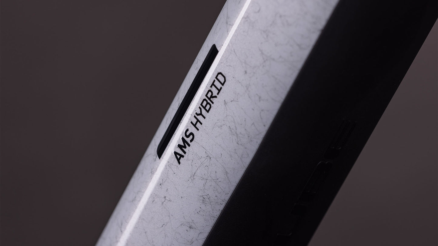 Nahaufnahme des Cube AMS Hybrid ONE44 C:68X SUPER TM 400X von CUBE mit dem Schriftzug "AMS HYBRID" auf einem schlanken Carbonrahmen. Das Lichtspiel hebt die fortschrittlichen Eigenschaften des All-Mountain-Bikes vor einem dunklen, schlichten Hintergrund hervor.