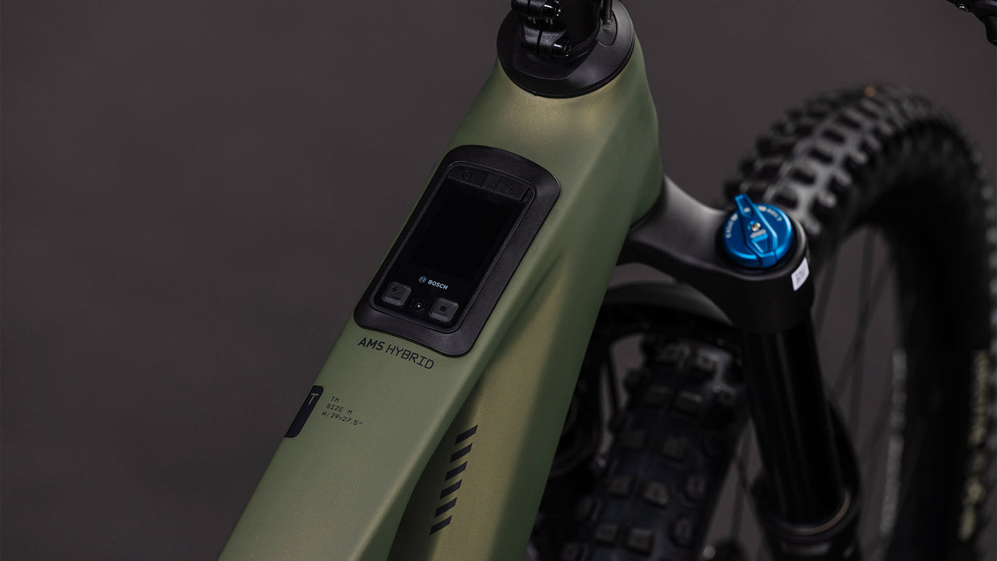 Nahaufnahme des CUBE AMS Hybrid 177 C:62 TM 600X reedgreen'n'matrix 2026: Man sieht den mattgrünen Carbonrahmen, die Digitalanzeige des Bosch CX-Motors am Lenker, die Federgabel und einen Teil des knorrigen Vorderreifens.
