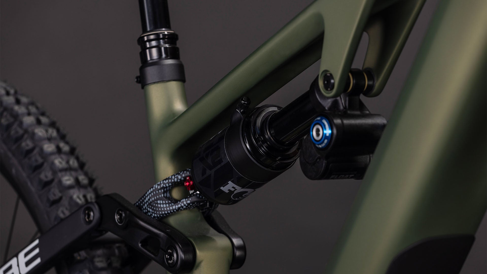 Nahaufnahme der Hinterradaufhängung eines CUBE AMS Hybrid 177 C:62 TM 600X reedgreen'n'matrix 2026 Mountainbikes, die den mattgrünen Carbonrahmen, den Stoßdämpfer und das Hinterrad zeigt. Schwarze Komponenten heben sich von einem dunklen Hintergrund ab.