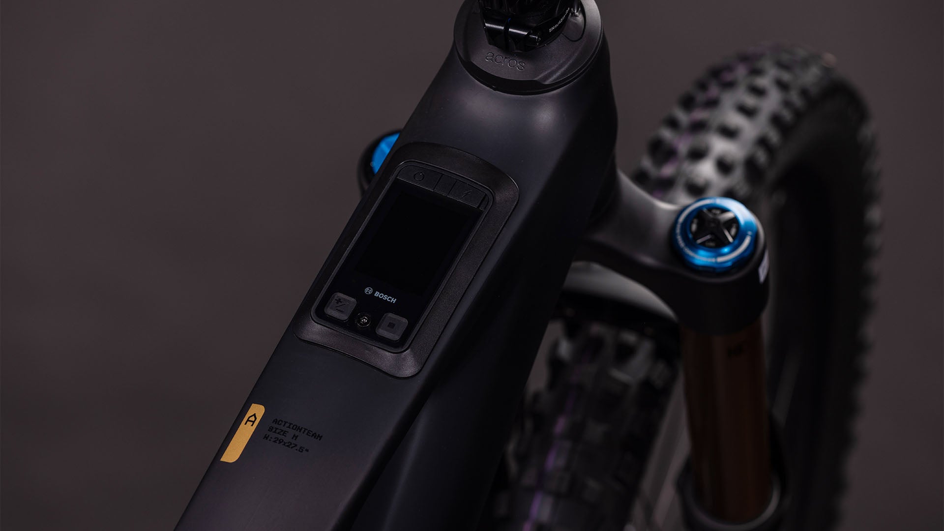 Nahaufnahme des Oberrohrs des CUBE AMS Hybrid 177 C:62 AT 600X carbon'n'gold 2026. Zu sehen sind das integrierte Bosch-Digitaldisplay, die Bosch CX-R Motorsteuerung und ein Teil der Vorderradaufhängung mit einem Stollenreifen im Hintergrund.