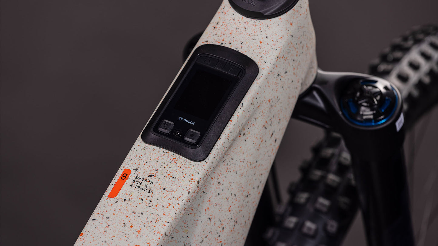 Nahaufnahme des CUBE AMS Hybrid 177 C:62 SUPER TM 600X (reedbeigegrit'n'orange, 2026) mit Bosch-Display, Bedientasten, Bosch CX-R-Motor, Fox Podium Federgabel, gesprenkeltem, grauweißem Rahmen, Warnaufkleber und Noppenreifen.
