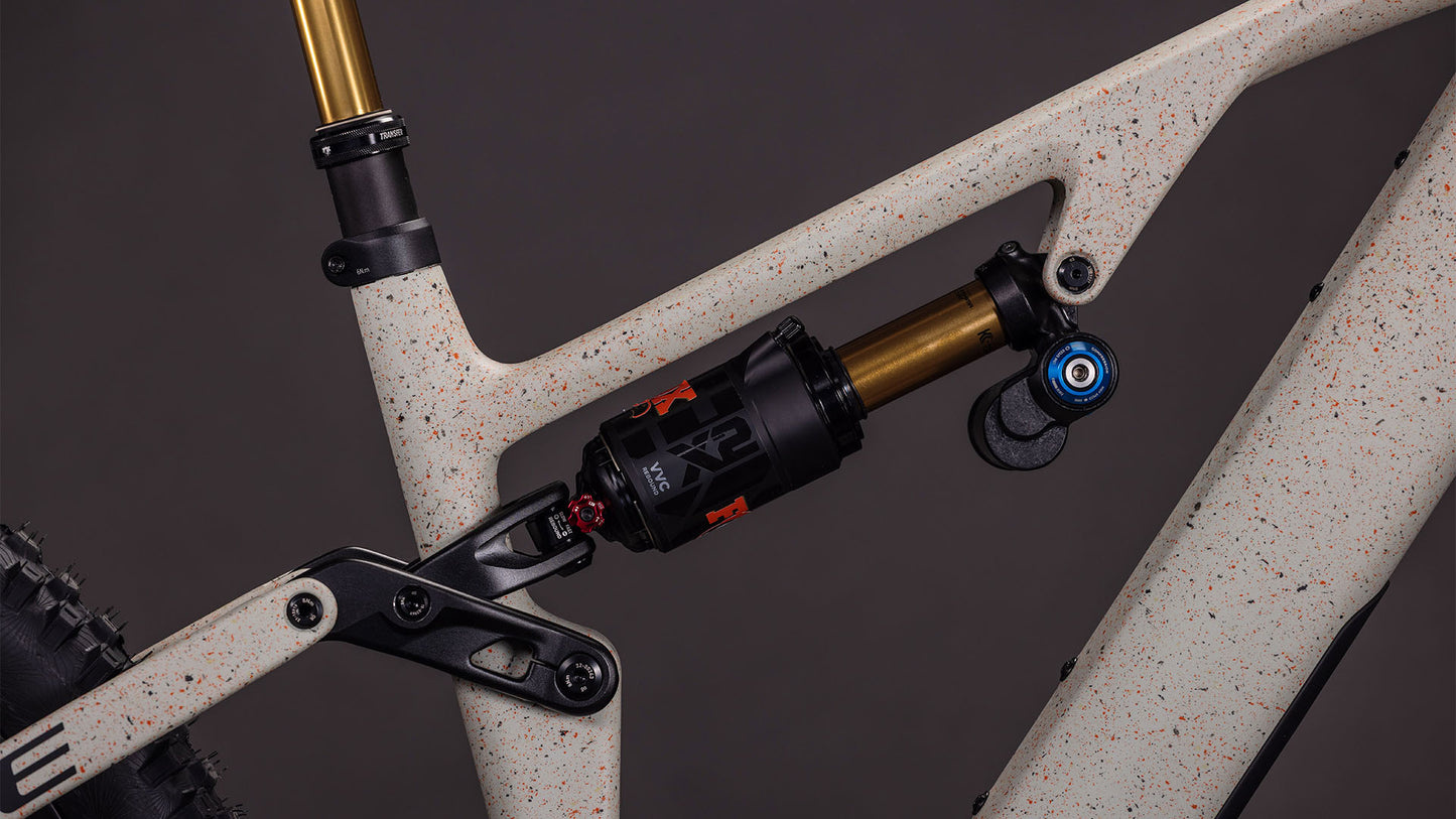 Nahaufnahme der CUBE AMS Hybrid 177 C:62 SUPER TM 600X Hinterradaufhängung, die einen schwarz-goldenen Stoßdämpfer auf einem gesprenkelten schilf-beigegrit'n'orangefarbenen Rahmen mit Hinterradreifen zeigt - ausgestattet mit einem Sram XX Eagle Getriebe.