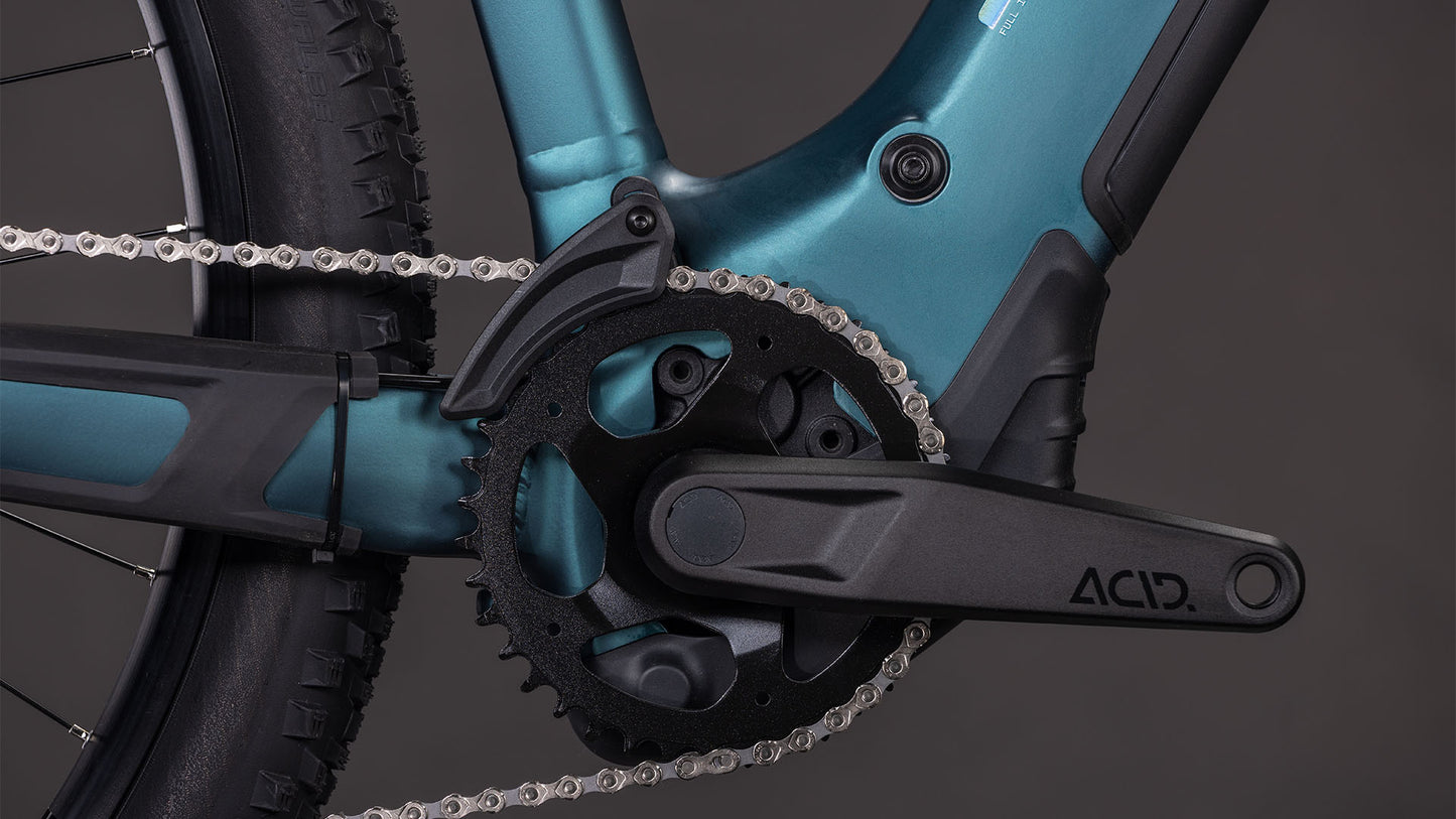 Nahaufnahme des CUBE-Kurbelarms und des Kettenblatts an einem Cube Reaction Hybrid ONE 800 royalgreen'n'prism Trapeze 2026 E-Bike-Rahmen, mit einem Teil des Hinterradreifens und des Bosch-Motors, vor einem grauen Hintergrund.
