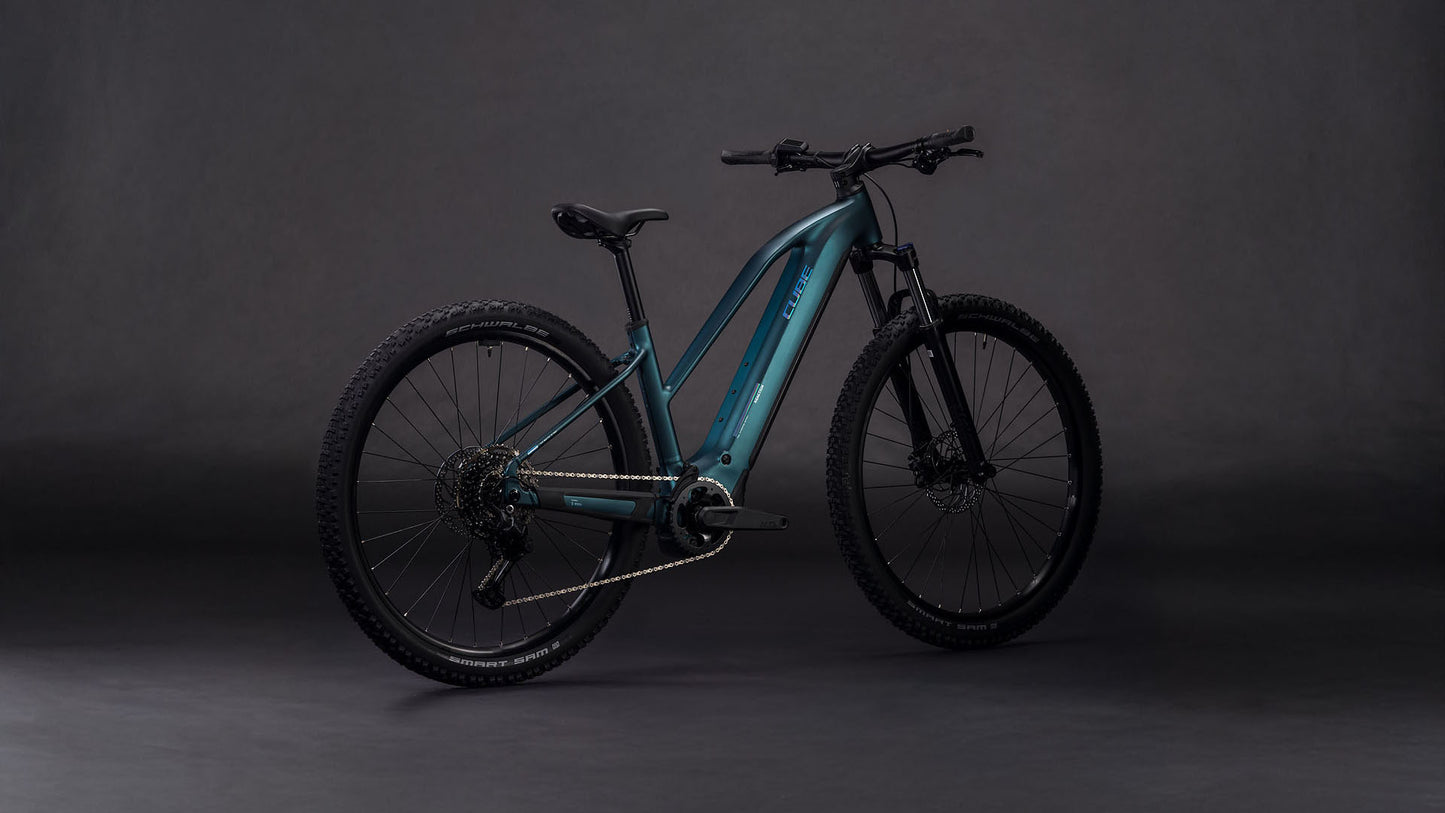 Das CUBE Reaction Hybrid ONE 800 royalgreen'n'prism Trapeze 2026 E-Bike verfügt über einen Bosch-Motor, breite Reifen, Scheibenbremsen, einen stabilen Rahmen und einen schwarzen Sattel, der vor einem dunklen, nahtlosen Hintergrund steht.