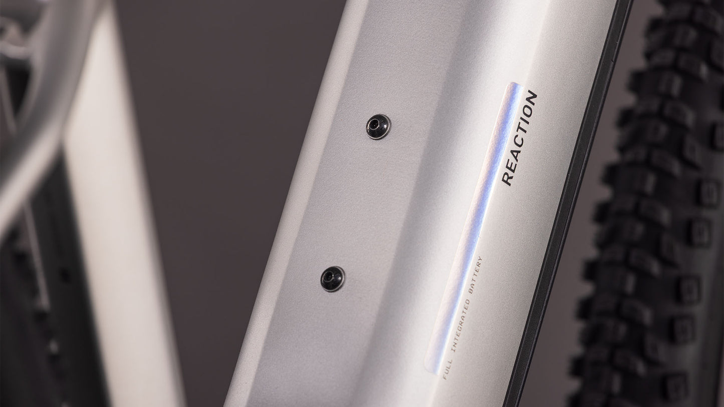 Nahaufnahme des CUBE Reaction Hybrid ONE 800 sleekgrey'n'prism 2026 Rahmens mit zwei Befestigungsschrauben und "REACTION" Aufschrift. Ein knorriger Reifen ist sichtbar und deutet auf den Bosch-Motor und den leistungsstarken 800-Wh-Akku hin.