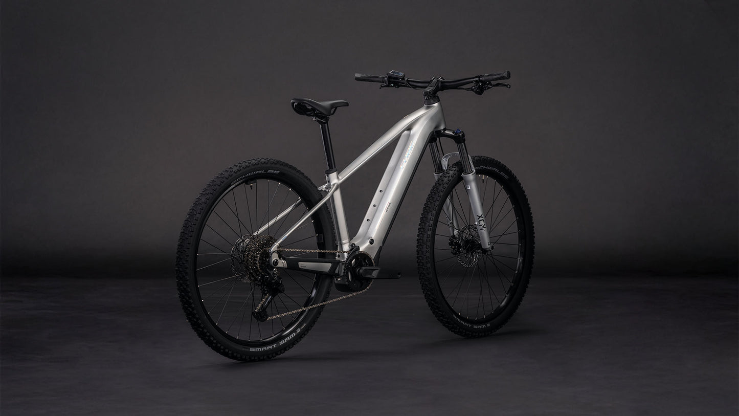 Das CUBE Reaction Hybrid ONE 800 sleekgrey'n'prism 2026 Mountainbike mit dicken Stollenreifen, mattsilbernem Rahmen und Bosch-Motor ist auf einem dunkelgrauen Hintergrund mit Front- und Seitenansicht abgebildet.