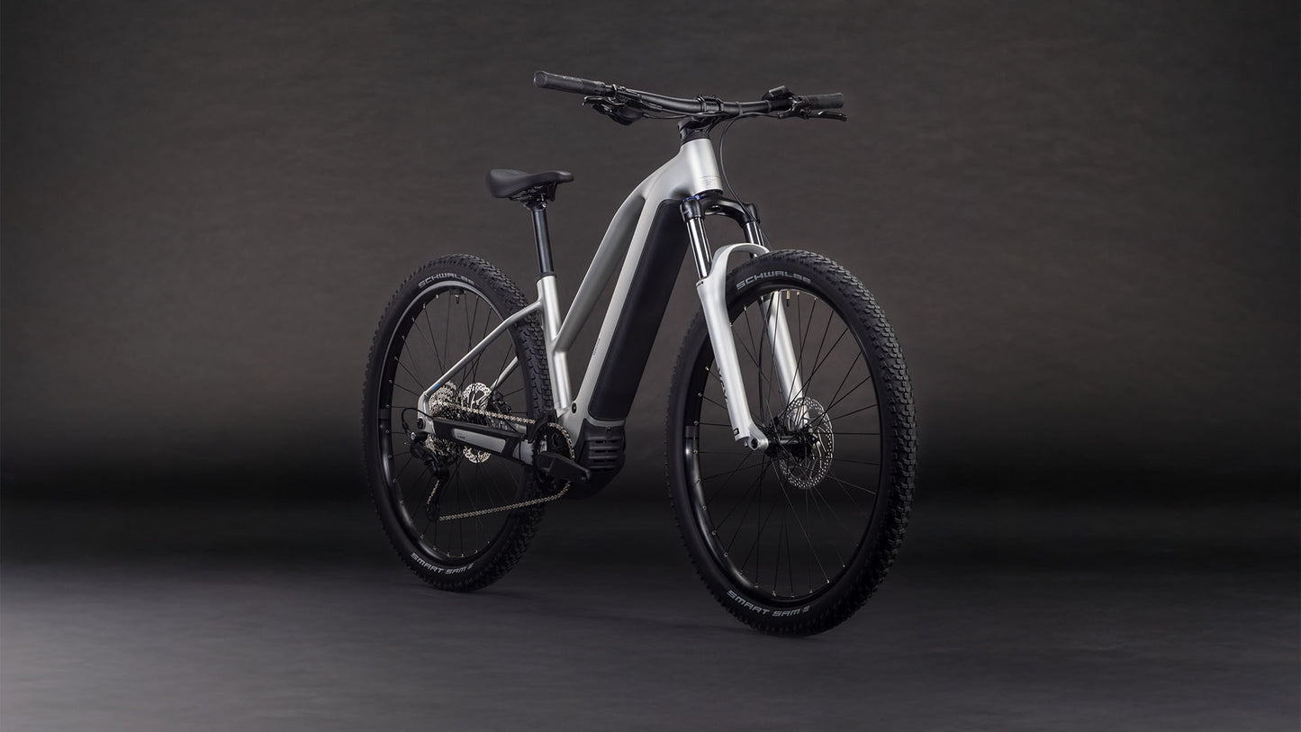Das CUBE Reaction Hybrid ONE 800 sleekgrey'n'prism Trapeze 2026 Elektrofahrrad mit mattsilbernem Rahmen, Bosch-Motor, dicken Reifen und geradem Lenker wird vor einem dunklen Hintergrund mit Farbverlauf gezeigt.