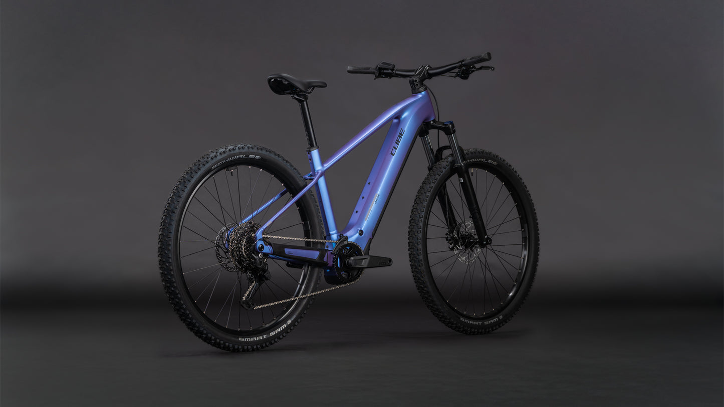 Das CUBE Reaction Hybrid ONE 800 blueiris'n'reflect 2026 Elektro-Mountainbike mit Bosch-Motor, metallisch blauem Rahmen, dicken Stollenreifen und eingebautem 800-Wh-Akku steht vor einem dunklen Hintergrund.