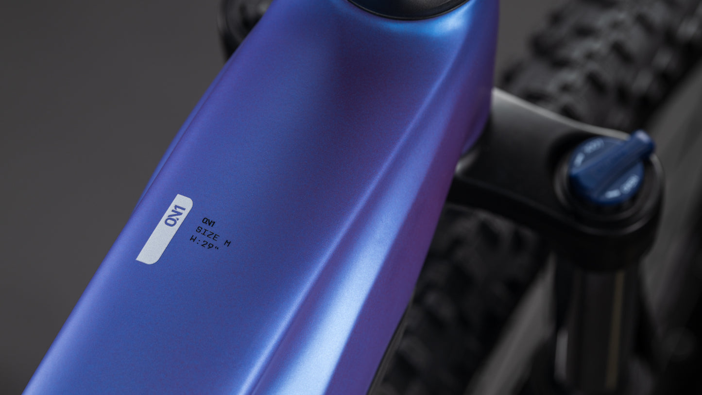 Nahaufnahme des CUBE Reaction Hybrid ONE 800 blueiris'n'reflect Trapeze 2026 Rahmens, die das metallisch blaue Oberrohr mit einem ON SIZE: M Aufkleber und einen Teil des Hinterreifens zeigt. Der Hintergrund ist unscharf.