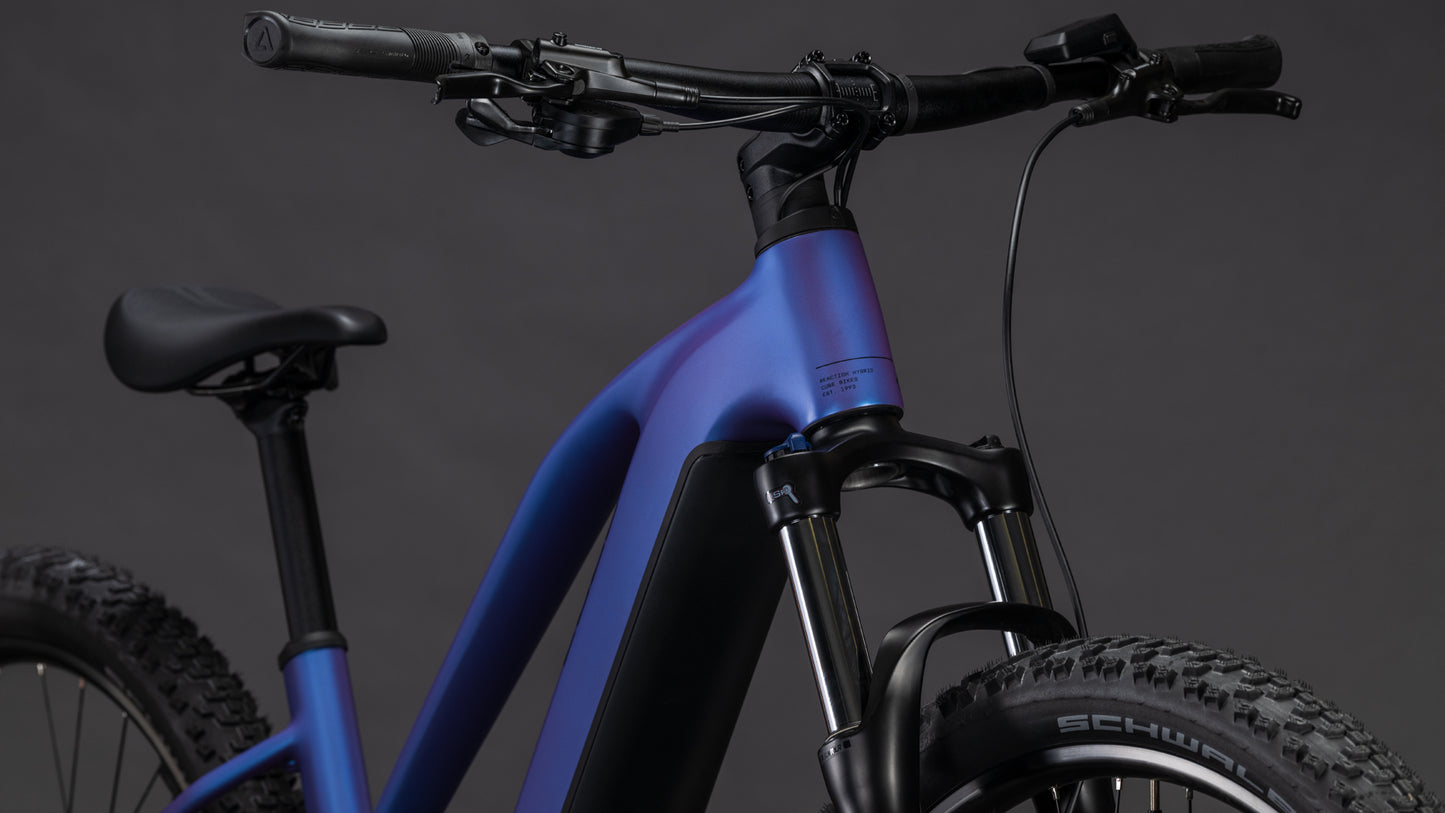 Nahaufnahme des CUBE Cube Reaction Hybrid ONE 800 blueiris'n'reflect Trapeze 2026 E-Bike mit mattblauem und schwarzem Rahmen, Bosch-Motor, Federgabel, breiten Schwalbe-Noppenreifen, flachem Lenker und dunklem Hintergrund.