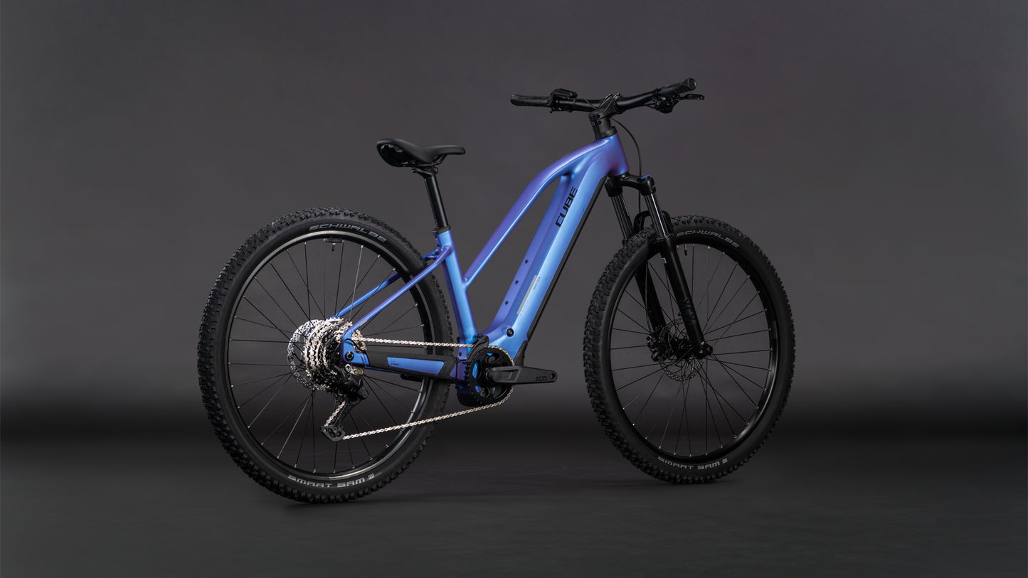 Das CUBE Reaction Hybrid ONE 800 blueiris'n'reflect Trapeze 2026 verfügt über einen metallisch blauen Rahmen, dicke Offroad-Reifen, einen Bosch-Motor, einen 800-Wh-Akku, eine Federgabel, Scheibenbremsen und einen aufrechten Sitz auf einem dunklen, neutralen Hintergrund.