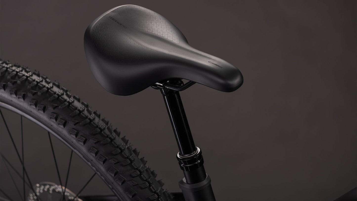 Nahaufnahme des schwarzen Sattels des CUBE Cube Reaction Hybrid Pro 800 blackline Trapeze 2026 auf der Sattelstütze, wobei ein Teil des Hinterrads und des Reifens des E-Mountainbikes vor einem dunklen Hintergrund zu sehen ist.