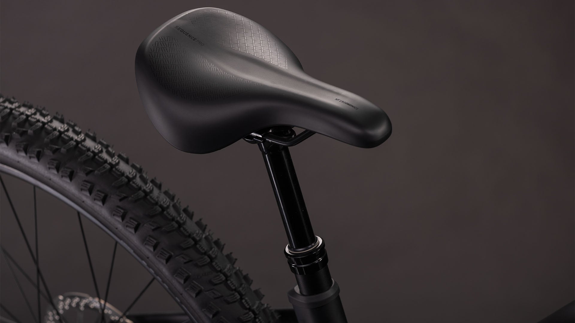 Nahaufnahme des schwarzen Sattels des CUBE Cube Reaction Hybrid Pro 800 blackline Trapeze 2026 auf der Sattelstütze, wobei ein Teil des Hinterrads und des Reifens des E-Mountainbikes vor einem dunklen Hintergrund zu sehen ist.