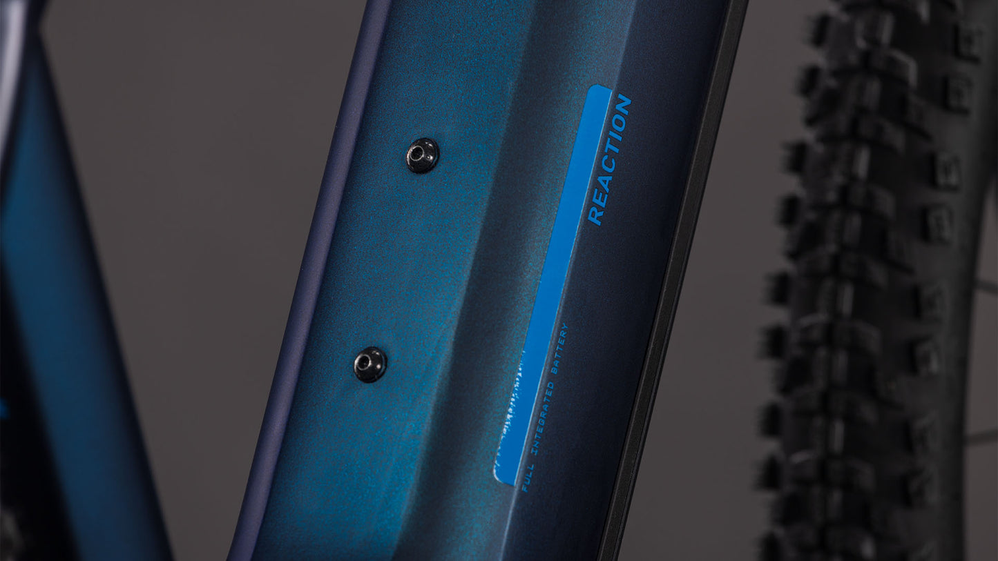 Nahaufnahme des Cube Reaction Hybrid Pro 800 nebular'n'dazzle 2026 Rahmens von CUBE mit zwei Befestigungsschrauben und REACTION Schriftzug in Blau. Im Hintergrund ist eine knorrige Fahrradreifenkante zu sehen.