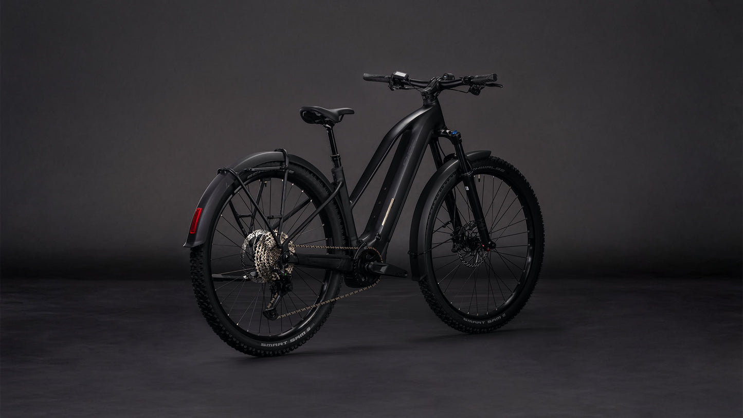 Das CUBE Reaction Hybrid Pro 800 FE blackline Trapeze 2026, ein mattschwarzes E-Bike mit dicken Reifen, Hinterradschutzblech und Scheibenbremsen, ausgestattet mit einem Bosch CX Motor, wird in einem Studio mit dunklem Hintergrund ausgestellt und von der Seite und von hinten gezeigt.